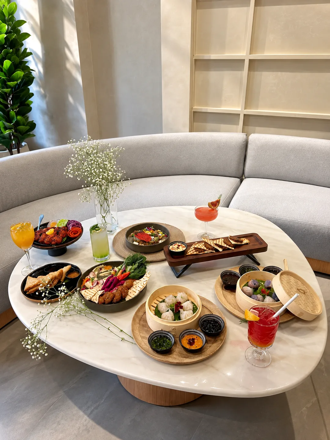 Elevated view of a sweeping crescent light grey banquette, oval marble table laden with culinary delicacies, and lush indoor greenery.