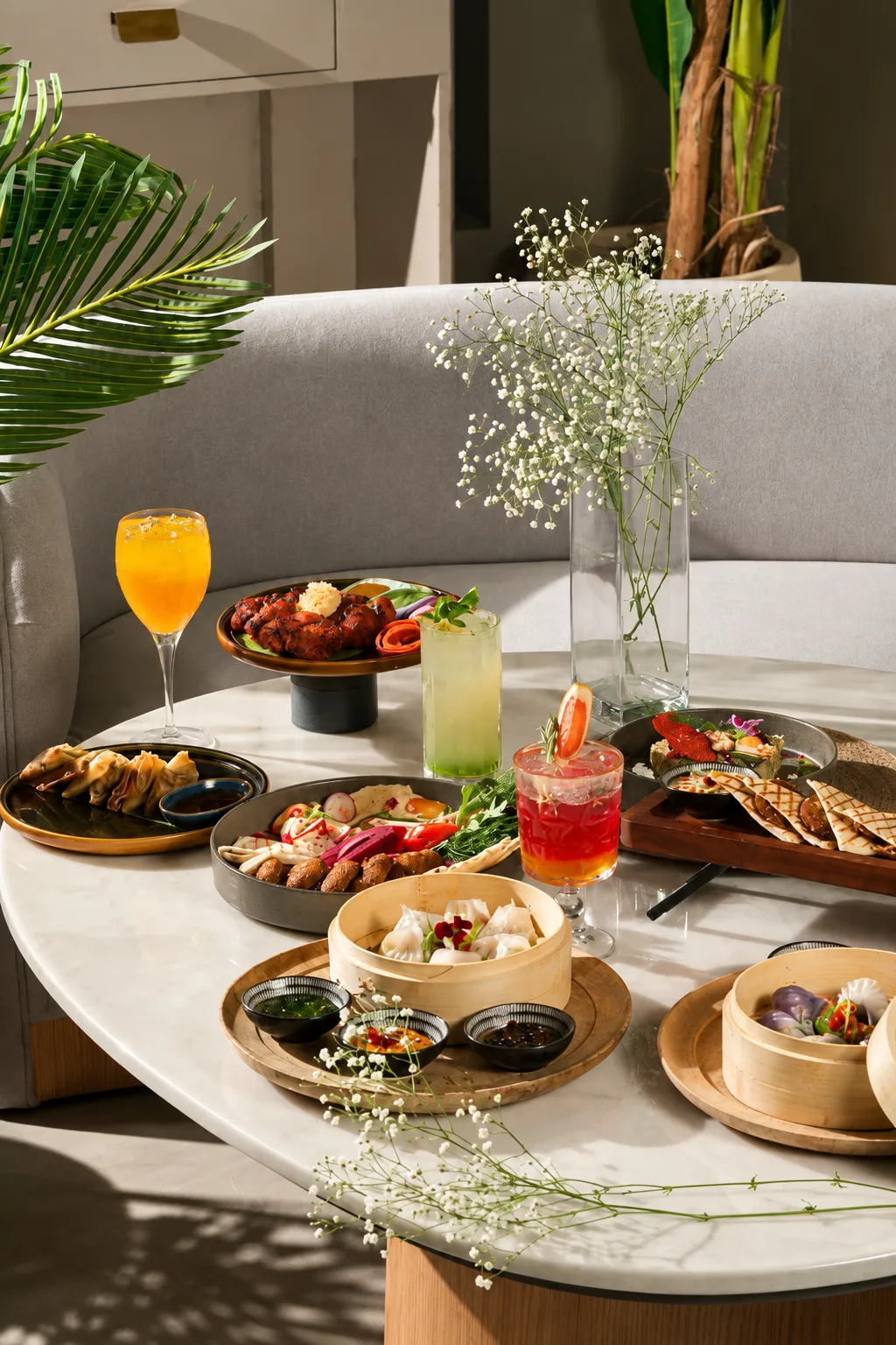 Sunlit marble dining table set with dimsums in bamboo steamers, artisan craft cocktails, mezze platter, and delicate baby’s breath flowers beside a plush banquette.