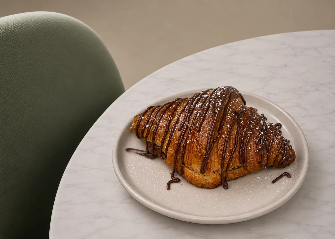 Golden flaky artisan croissant drizzled with Belgian chocolate ganache on a warm stone plate beside a fluted sage green velvet chair.