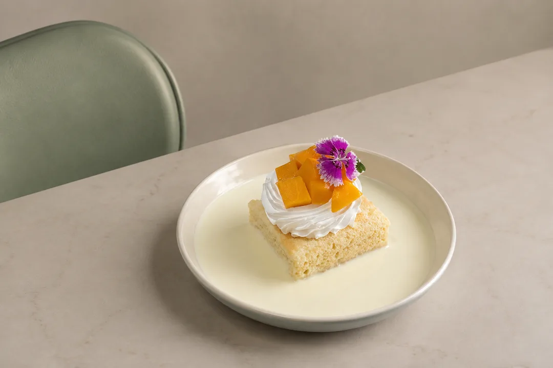 Saffron mango soaked sponge cake topped with delicate piped chantilly cream and an edible purple flower on an ivory ceramic plate.